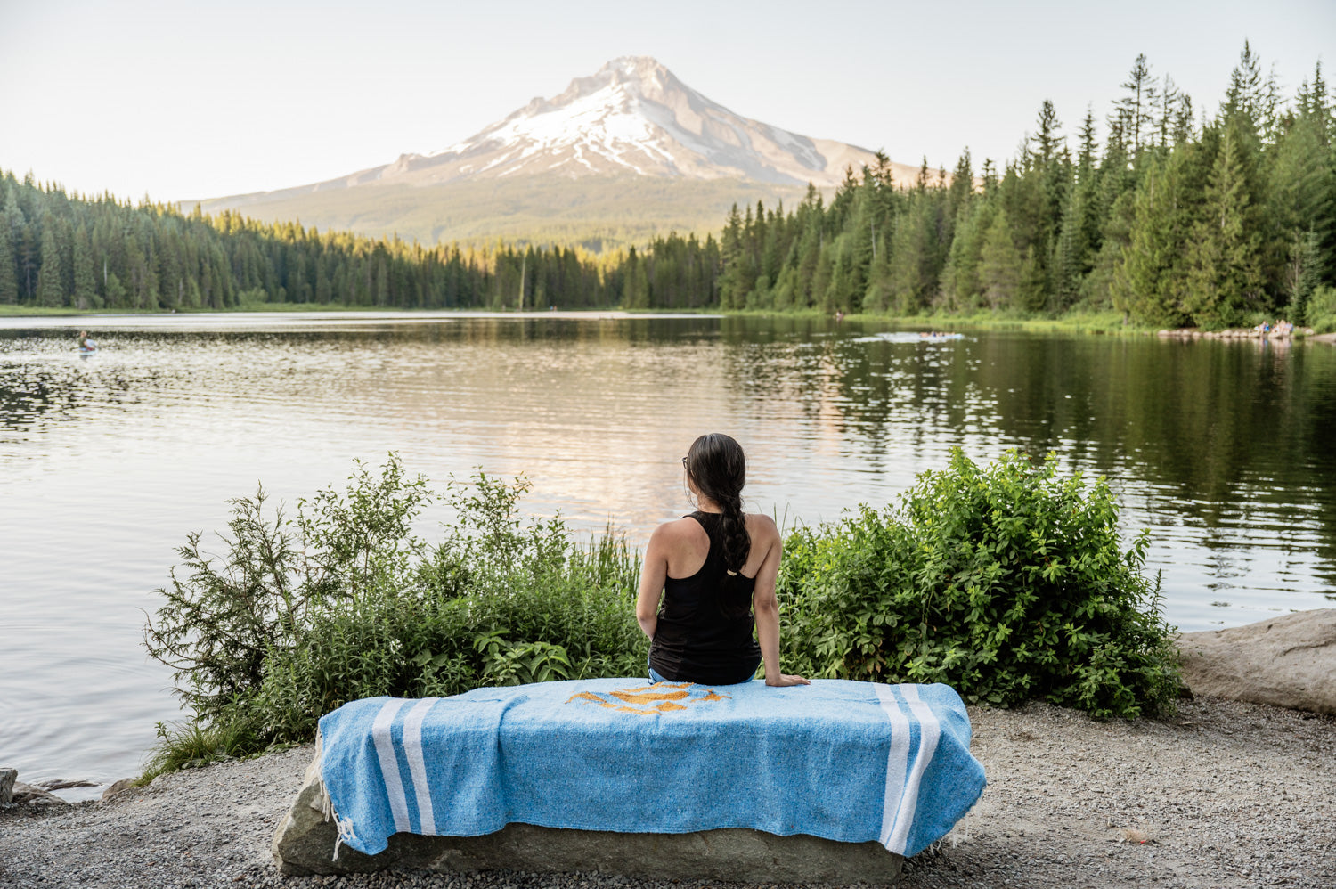 Woman sitting on blue blanket by a lake and mountain at sunset