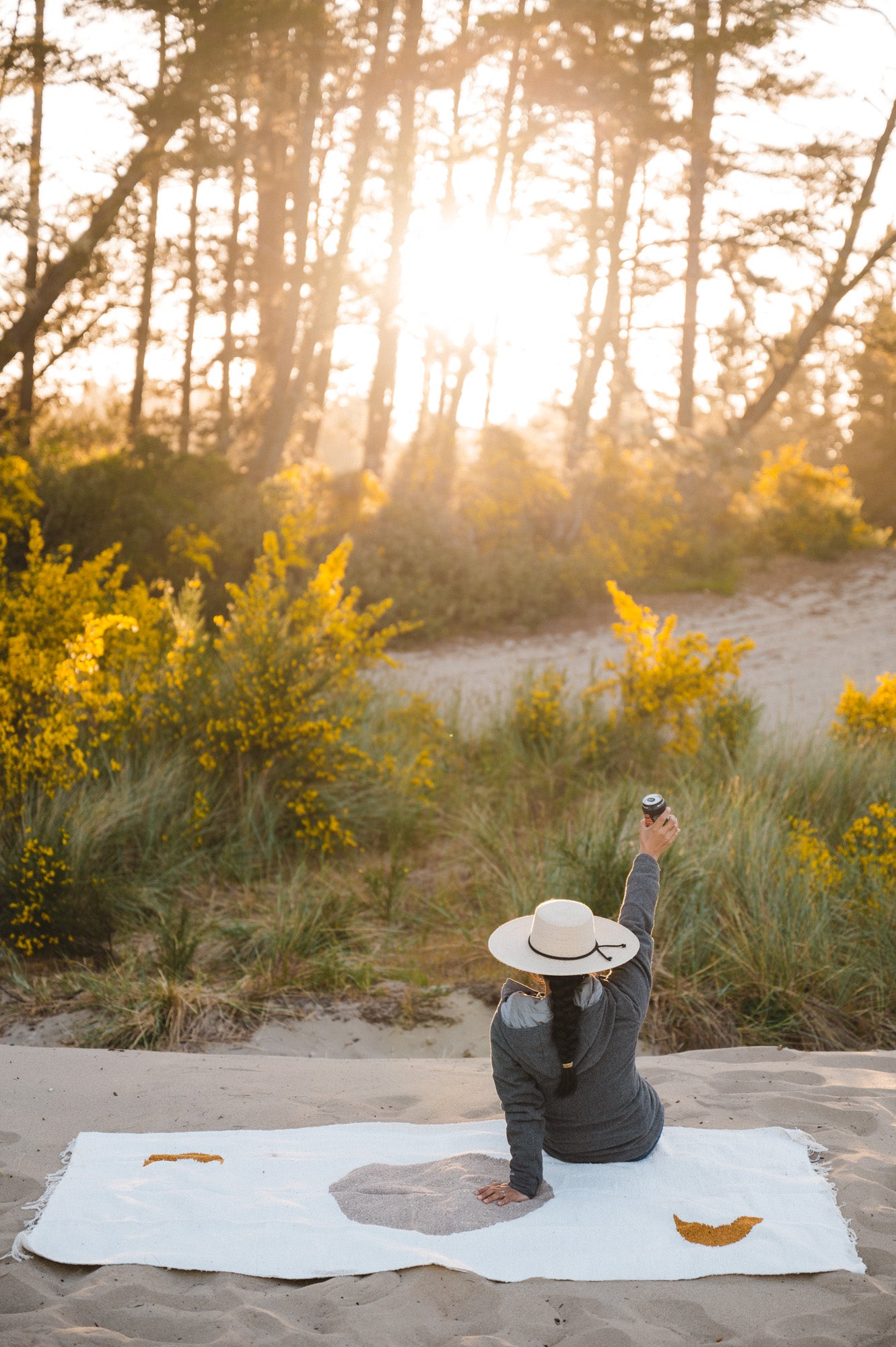 Woman holding a beer sitting on cream blanket in the sand at sunset