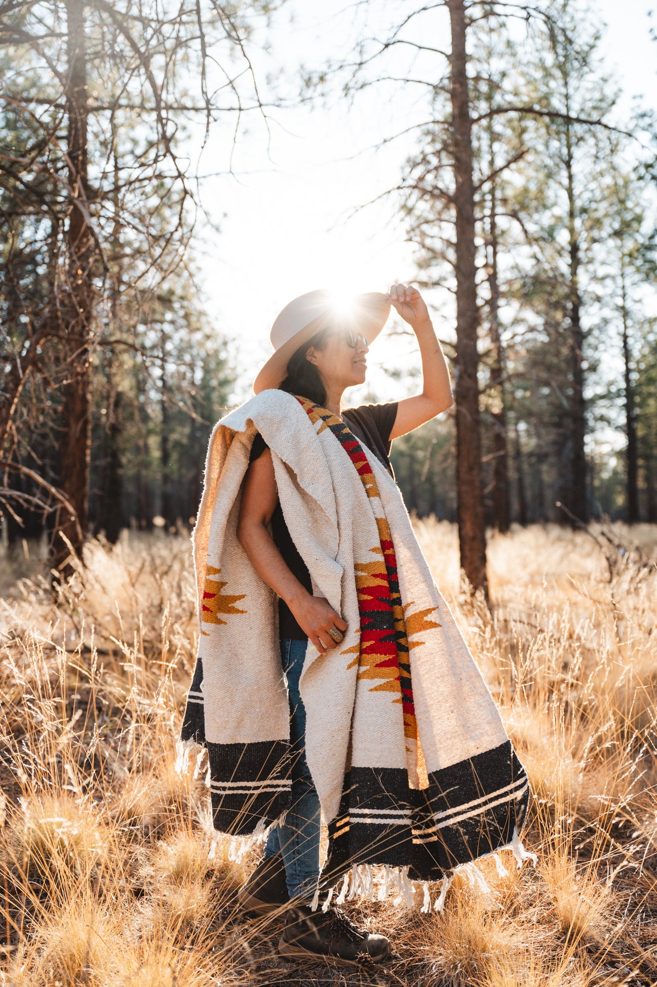 Woman wearing cowboy hat standing in a dry grassy field and forest at sunset wearing a southwestern diamond blanket over her shoulder