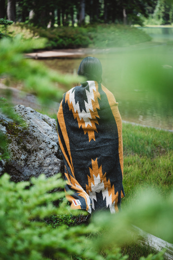 Girl wearing black and yellow zapotec handwoven mexican blanket in front of alpine lake