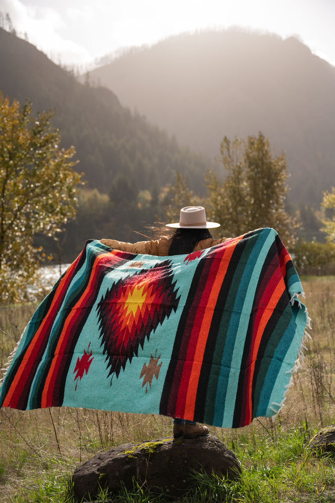 Woman wearing a cowboy hat holding a teal and red handwoven mexican diamond blanket in front of sunset in the mountains