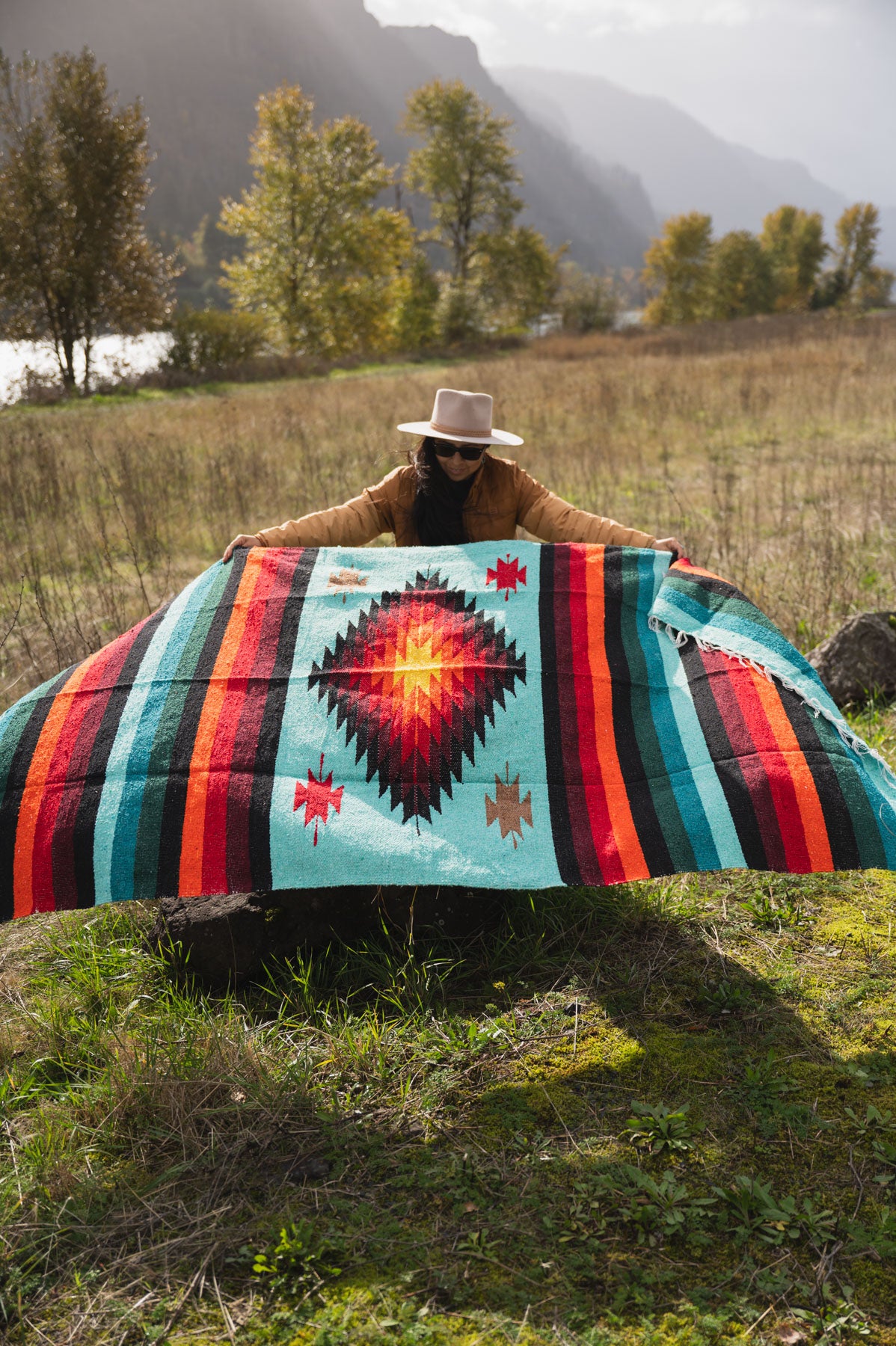 Woman wearing a cowboy hat holding a teal and red handwoven mexican diamond blanket in front of sunset in the mountains