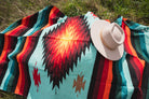 teal and red handwoven diamond mexican blanket laying on the ground with a cowboy hat on top of it. 