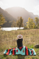 Woman wearing a cowboy hat sitting on a teal and red handwoven mexican diamond blanket in front of sunset in the mountains