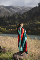 Woman wrapped in a a teal and red handwoven mexican diamond blanket in front of sunset in the mountains