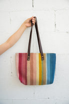 Rainbow colored striped Mexican handbag being held against a white brick wall by an arm