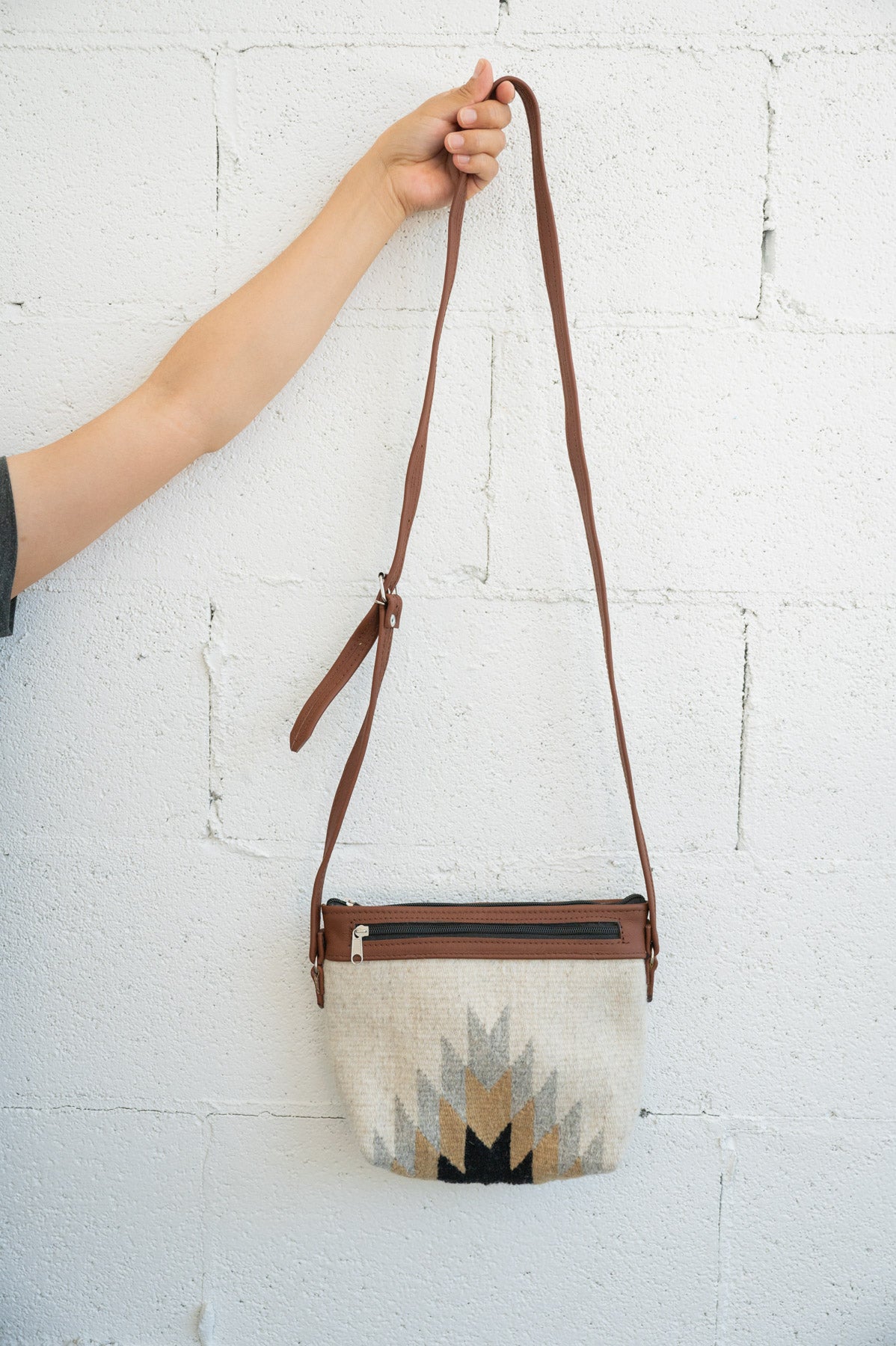 Cream, grey and gold Mexican shoulder bag being held against a white brick wall by an arm