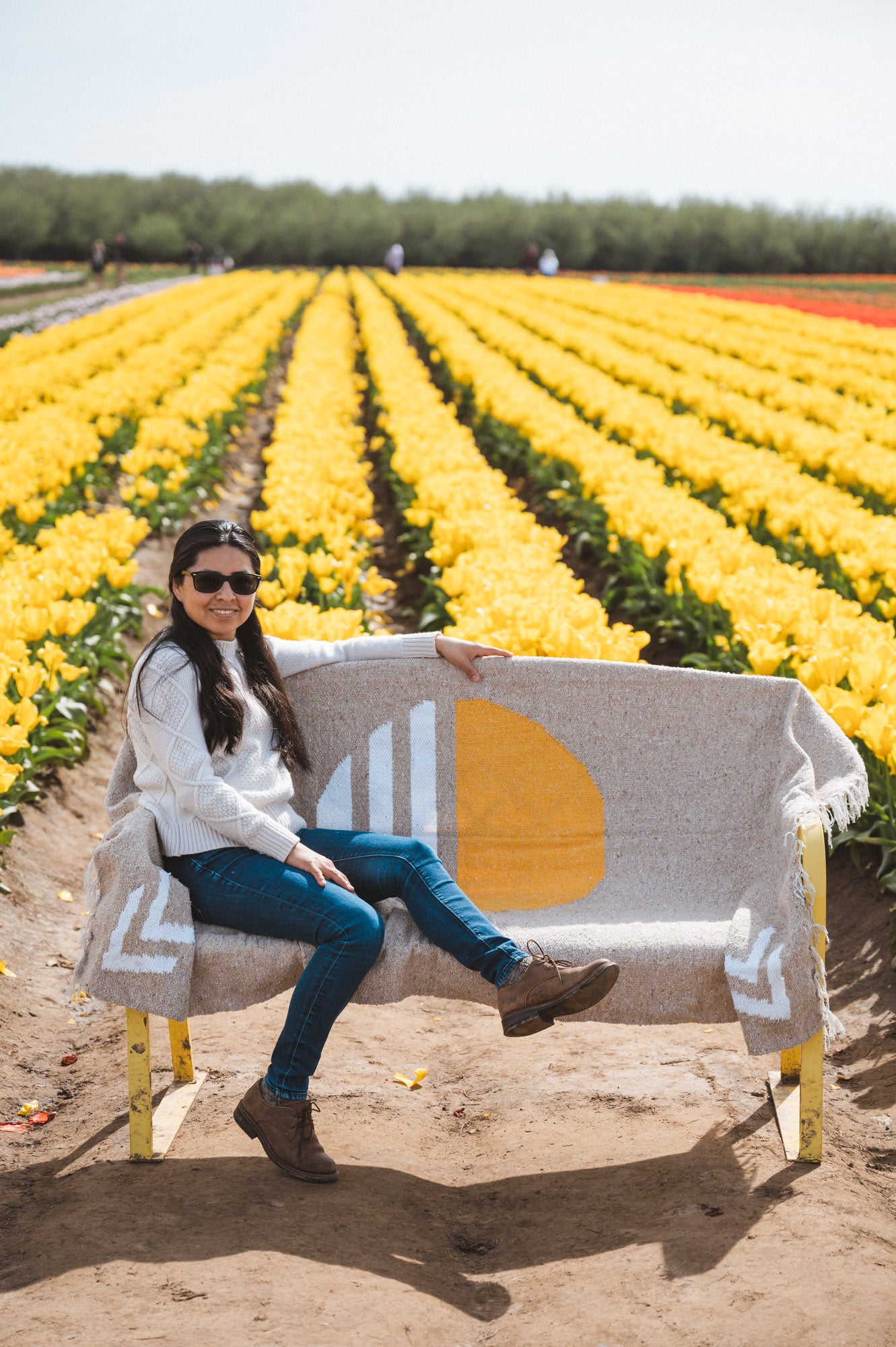 lady sitting on bench that are covered with tan sun blanket, in the back there is a view of yellow flower farm