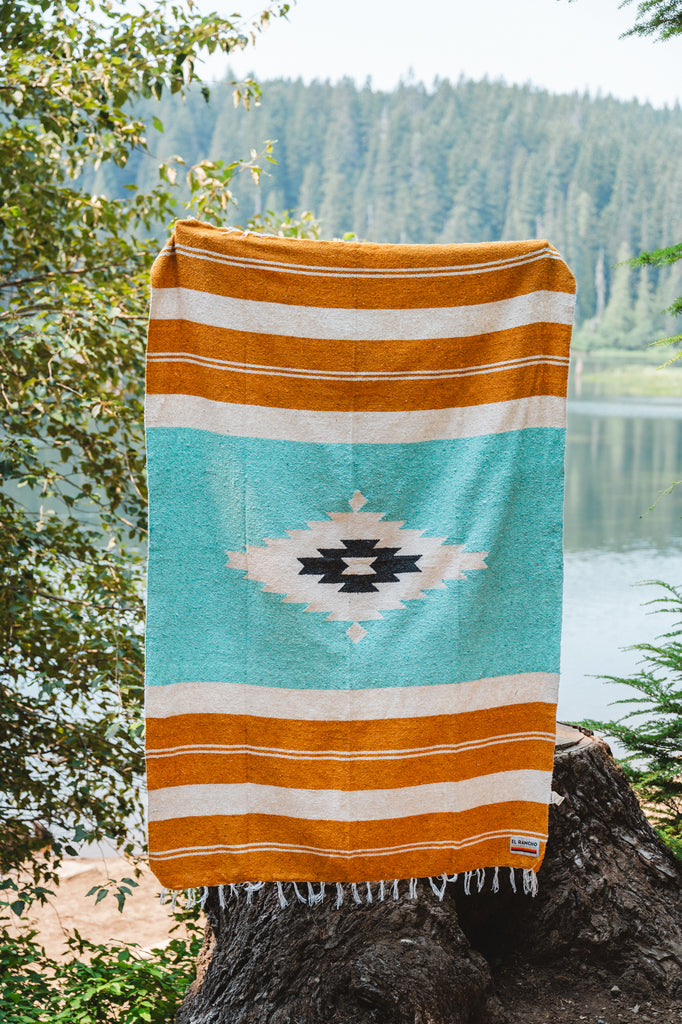 turquoise and gold diamond zapotec blanket being held up in front of a mountain lake